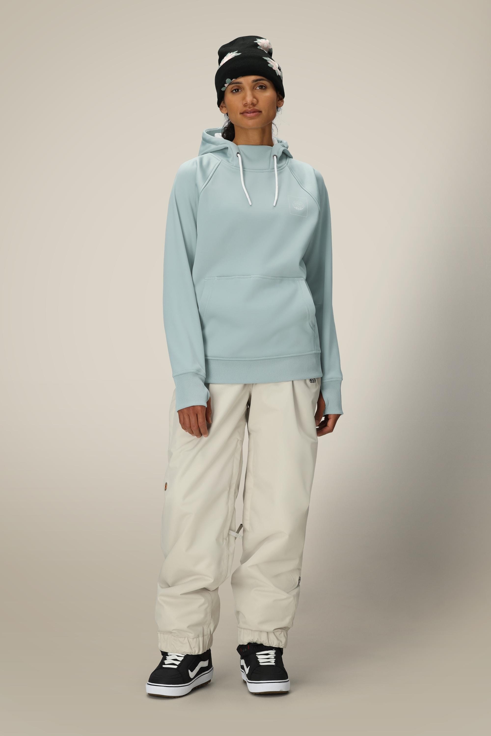 A person stands facing forward against a plain, light background, wearing the 686 Womens Bonded Fleece Pullover Hoody in light blue, off-white baggy pants, black Vans sneakers, and a black beanie with white accents.