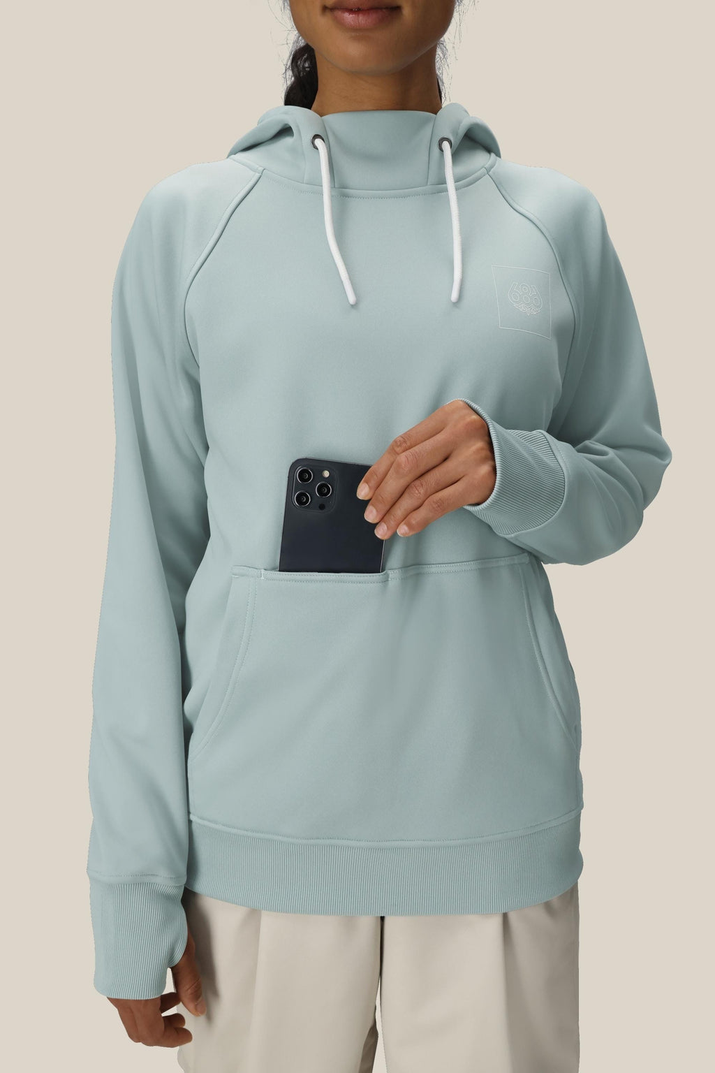 A woman in the 686 Womens Bonded Fleece Pullover Hoody, featuring a soft fleece lining and white drawstrings, places a black smartphone in the kangaroo pocket. She is also wearing light-colored pants; her face is partially visible.