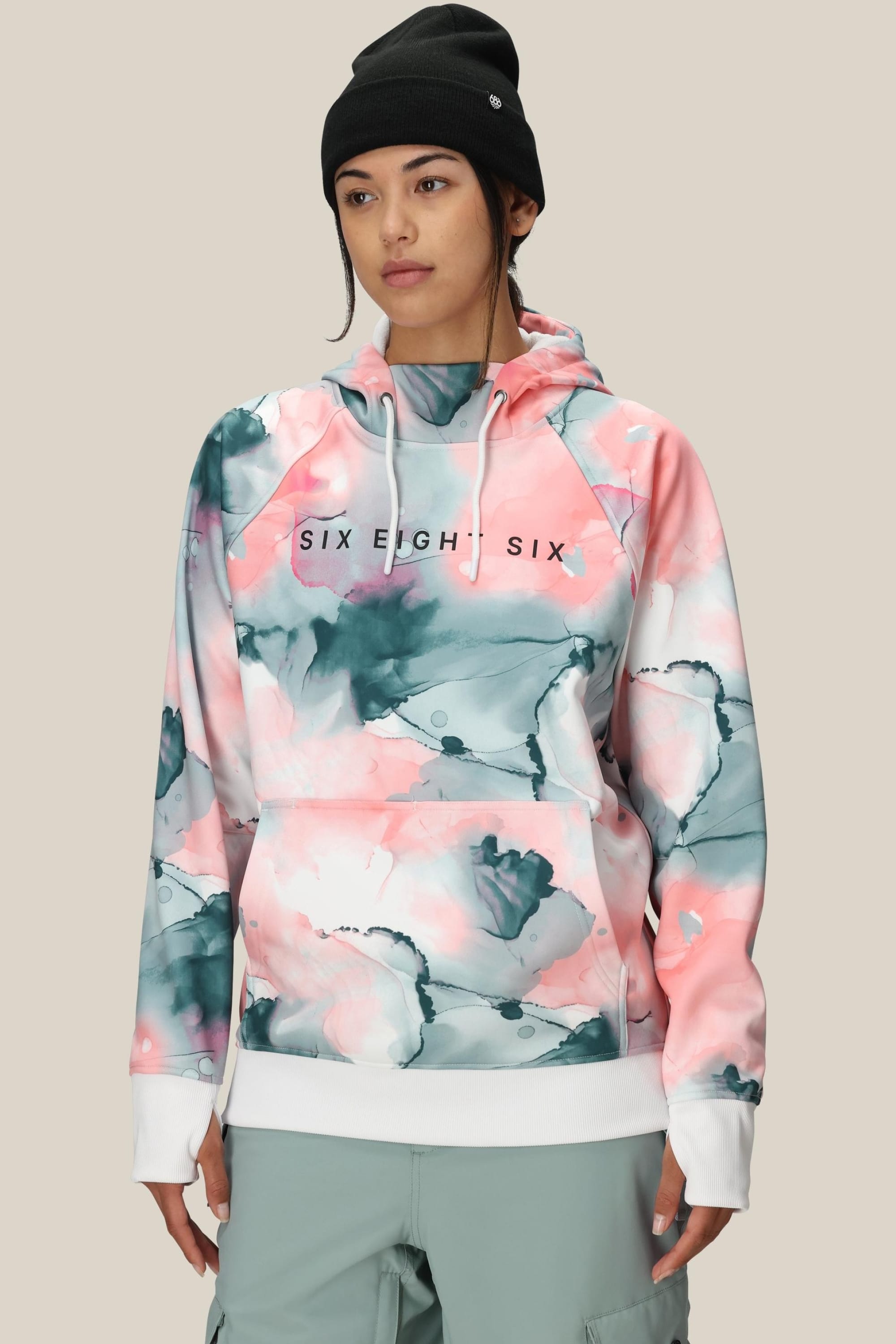 A person wears the 686 Womens Bonded Fleece Pullover Hoody—a fleece-lined, watercolor-patterned hoodie with SIX EIGHT SIX on the front—paired with a black beanie, standing against a plain beige background.