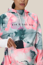 A woman wears the 686 Womens Bonded Fleece Pullover Hoody in pink, white, and gray tie-dye with SIX EIGHT SIX on the front, placing a black smartphone into its pouch pocket. Her face is partially visible.