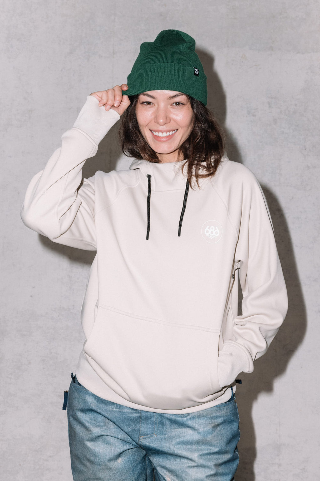 A smiling woman wears the 686 Womens Bonded Fleece Pullover Hoody, blue jeans, and a green knit beanie while standing against a light gray background, touching the brim of her beanie with one hand.