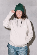 A smiling woman wears the 686 Womens Bonded Fleece Pullover Hoody, blue jeans, and a green knit beanie while standing against a light gray background, touching the brim of her beanie with one hand.