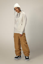 A person wearing a white beanie, the 686 Womens Bonded Fleece Pullover Hoody, loose brown pants, and black sneakers stands against a plain light background, looking at the camera with a relaxed expression.