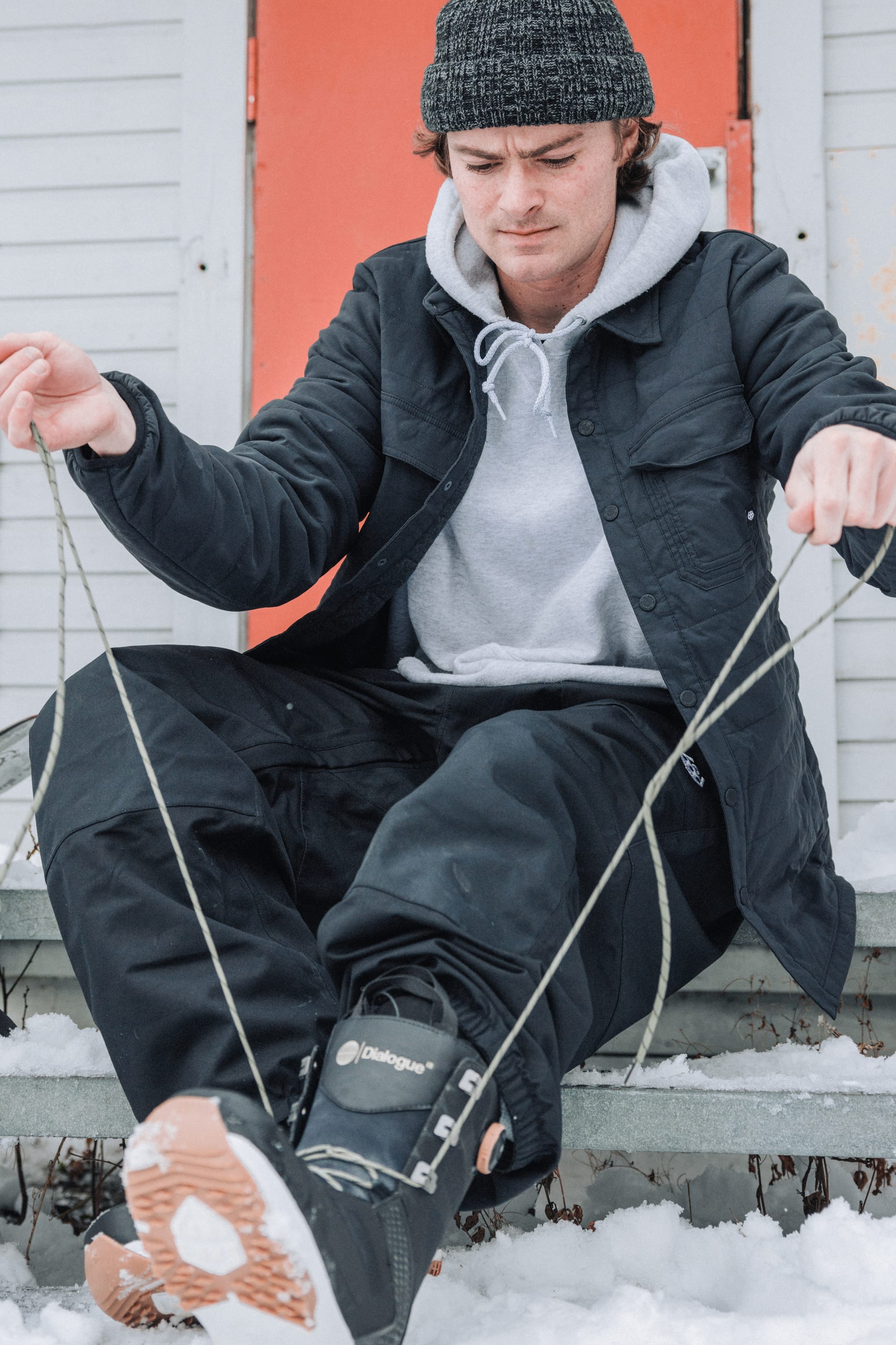 A person sits on snowy steps, focused as they lace up a black snowboarding boot, wearing the 686 Men's Thermaldry™ Snap-Up Shirt and beanie. A red door is visible behind them.