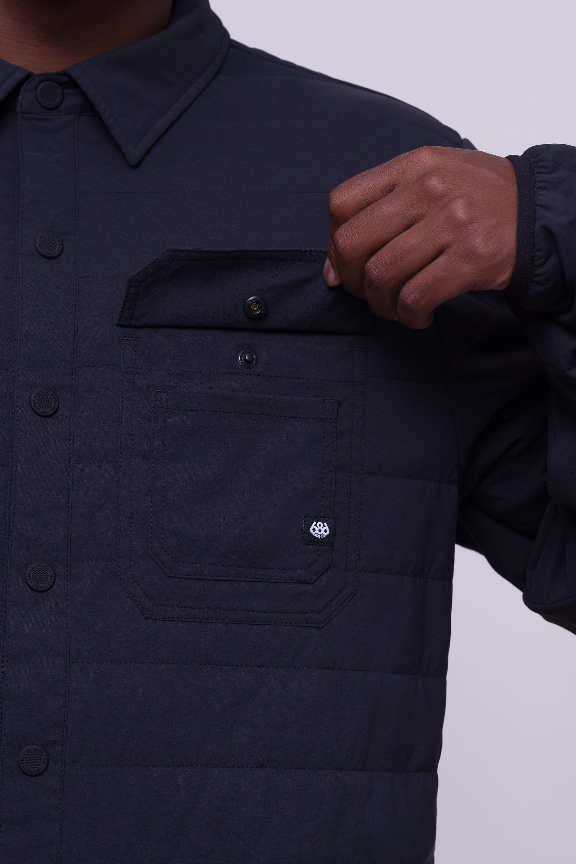 A person wears the 686 Men's Thermaldry™ Snap-Up Shirt, a dark quilted jacket with a chest pocket and button flap—ideal citywear by 686, blending urban style and subtle performance for any adventure.
