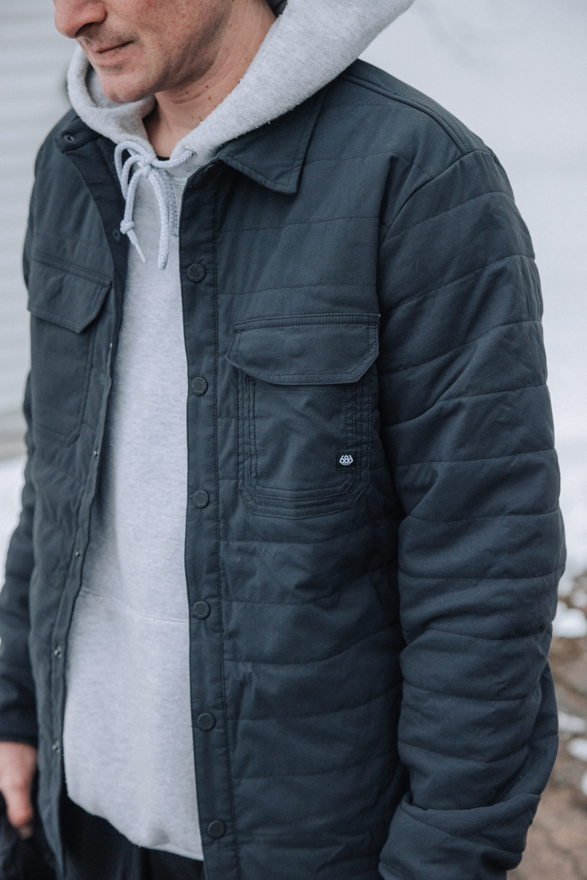 A man stands outdoors in winter, head slightly turned down and out of frame, wearing the 686 Men's Thermaldry™ Snap-Up Shirt by 686 layered under a dark blue quilted jacket, creating a stylish citywear look against a blurred background.