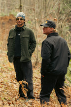 Two men stand in a forest clearing covered with dry leaves. One wears the 686 Men's Thermaldry™ Snap-Up Shirt with black pants, sunglasses, and a beanie; the other sports a black jacket, black pants, and a black cap.