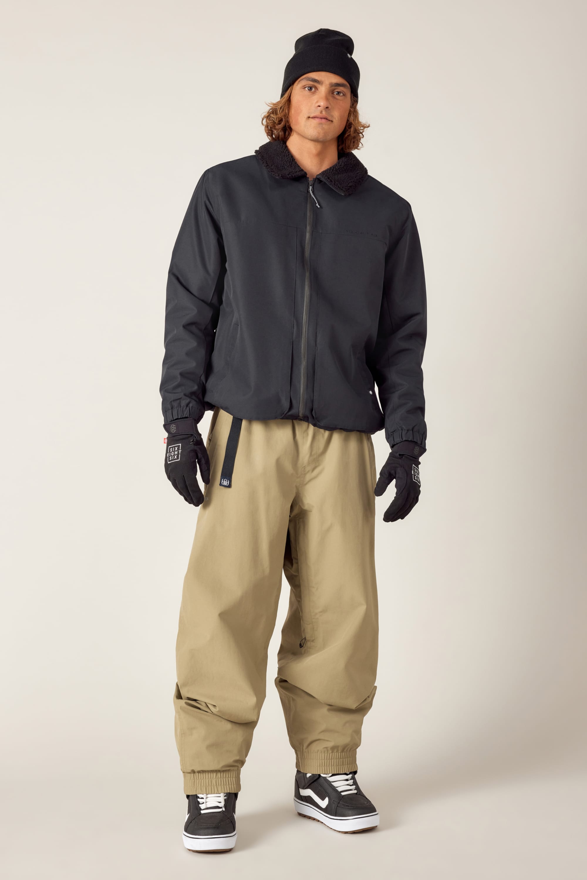A person stands facing forward against a plain, light background, wearing a black beanie, 686 Mens Caim Insulated Jacket by 686 with a fur collar, black gloves, loose tan pants, and black Vans sneakers.