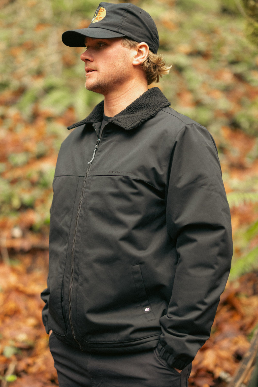 A person wearing the 686 Mens Caim Insulated Jacket and a black cap stands outdoors in a forest, surrounded by fallen leaves and greenery.