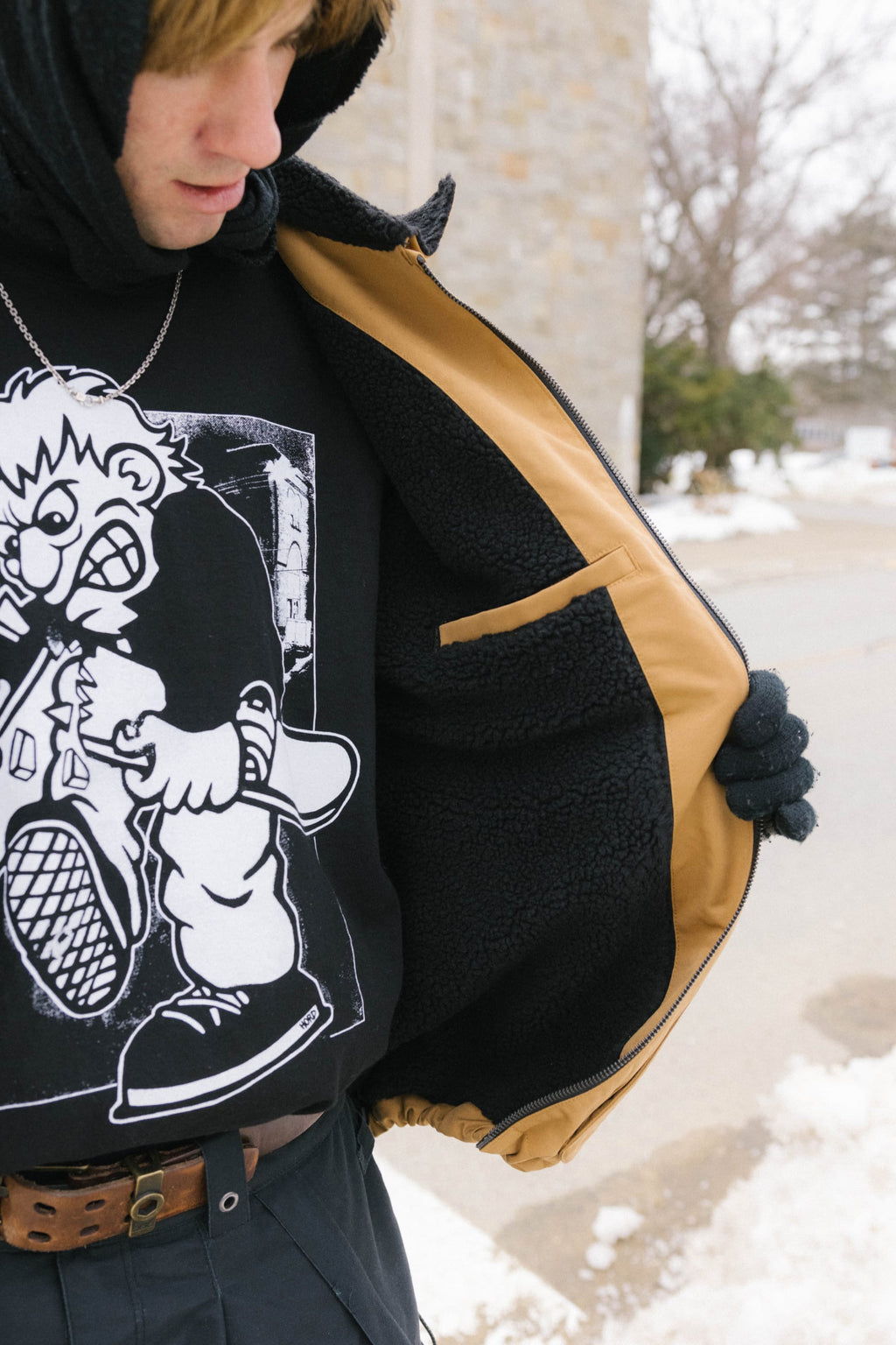 Outdoors in winter, a person opens a tan and black 686 Mens Caim Insulated Jacket to show a black sweatshirt with a large cartoon skateboarder graphic. Snow and trees appear in the background.
