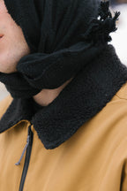 Close-up of a person wearing the 686 Mens Caim Insulated Jacket by 686, featuring a brown color, black fleece collar, and a black scarf wrapped around their head and neck. Only part of their face is visible with an out-of-focus background.
