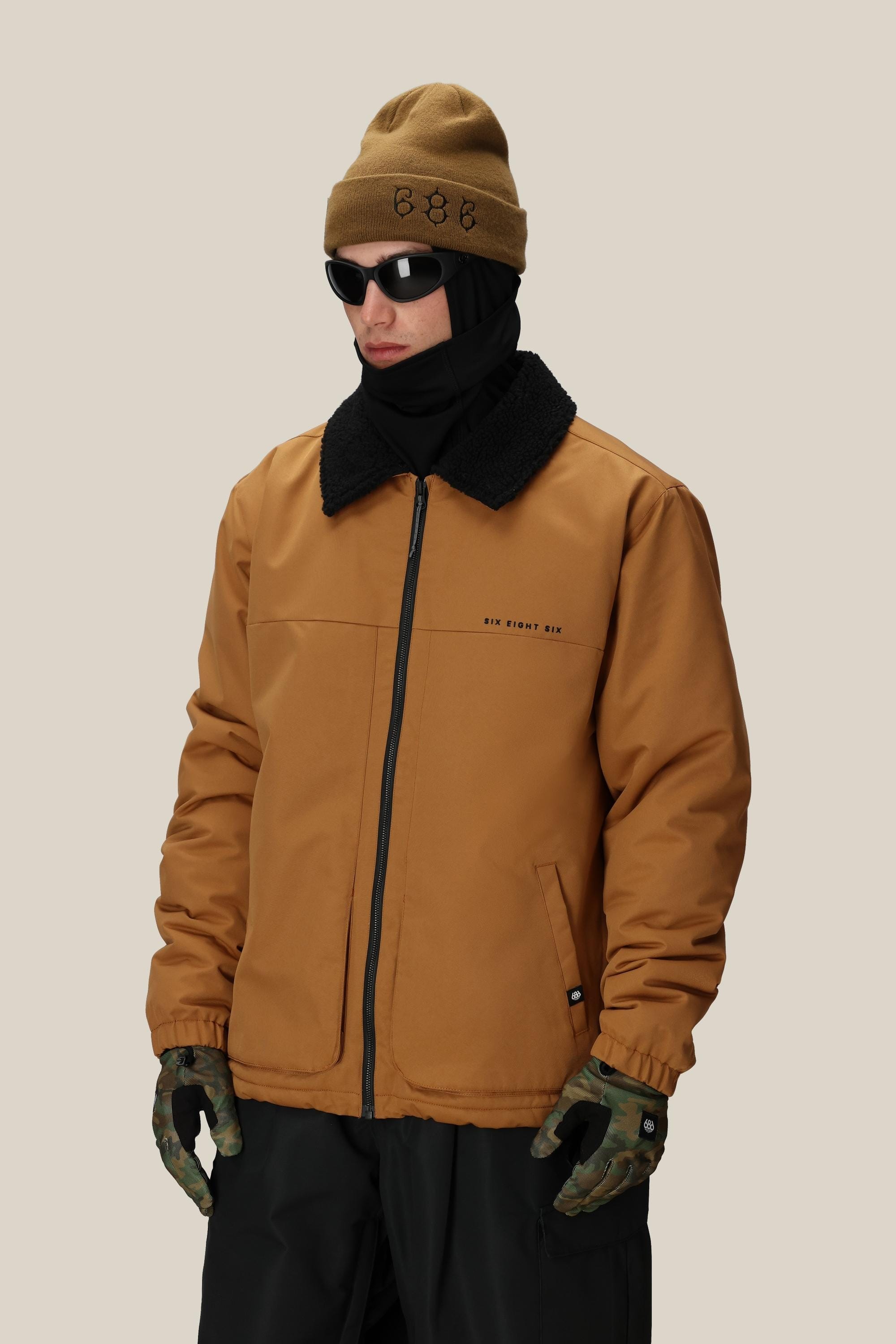 A person wearing the 686 Mens Caim Insulated Jacket by 686, paired with a brown beanie, black sunglasses, black scarf, and camouflage gloves stands against a plain beige background.