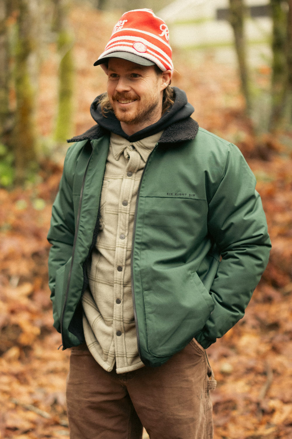 A man in a red hat and the 686 Mens Caim Insulated Jacket stands on a leafy forest path, smiling with his hands in his pockets.