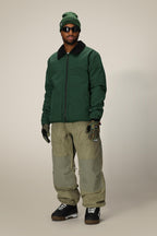 A person wearing the 686 Mens Caim Insulated Jacket stands facing forward against a plain light background, paired with olive green pants, black boots, gloves, a green beanie, and sunglasses.