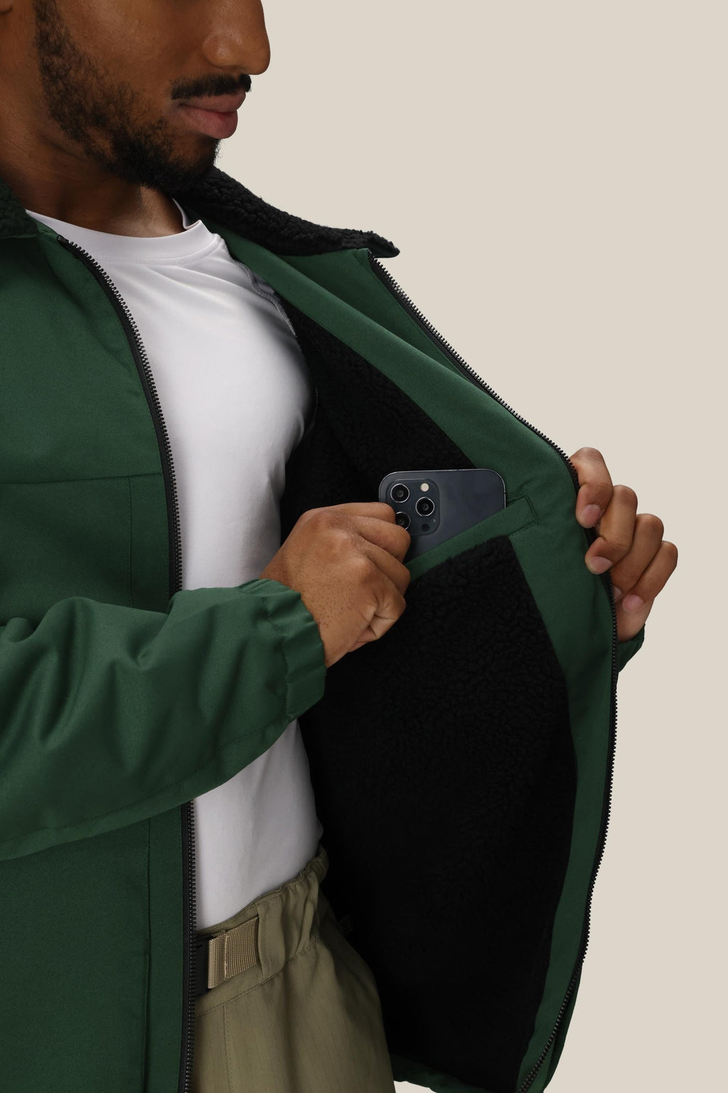 A person wearing the 686 Mens Caim Insulated Jacket partially unzipped, with a white shirt, places a dark smartphone into the inside pocket lined with black fleece. The 686 jacket’s interior pocket is visible.