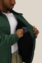 A person wearing the 686 Mens Caim Insulated Jacket partially unzipped, with a white shirt, places a dark smartphone into the inside pocket lined with black fleece. The 686 jacket’s interior pocket is visible.