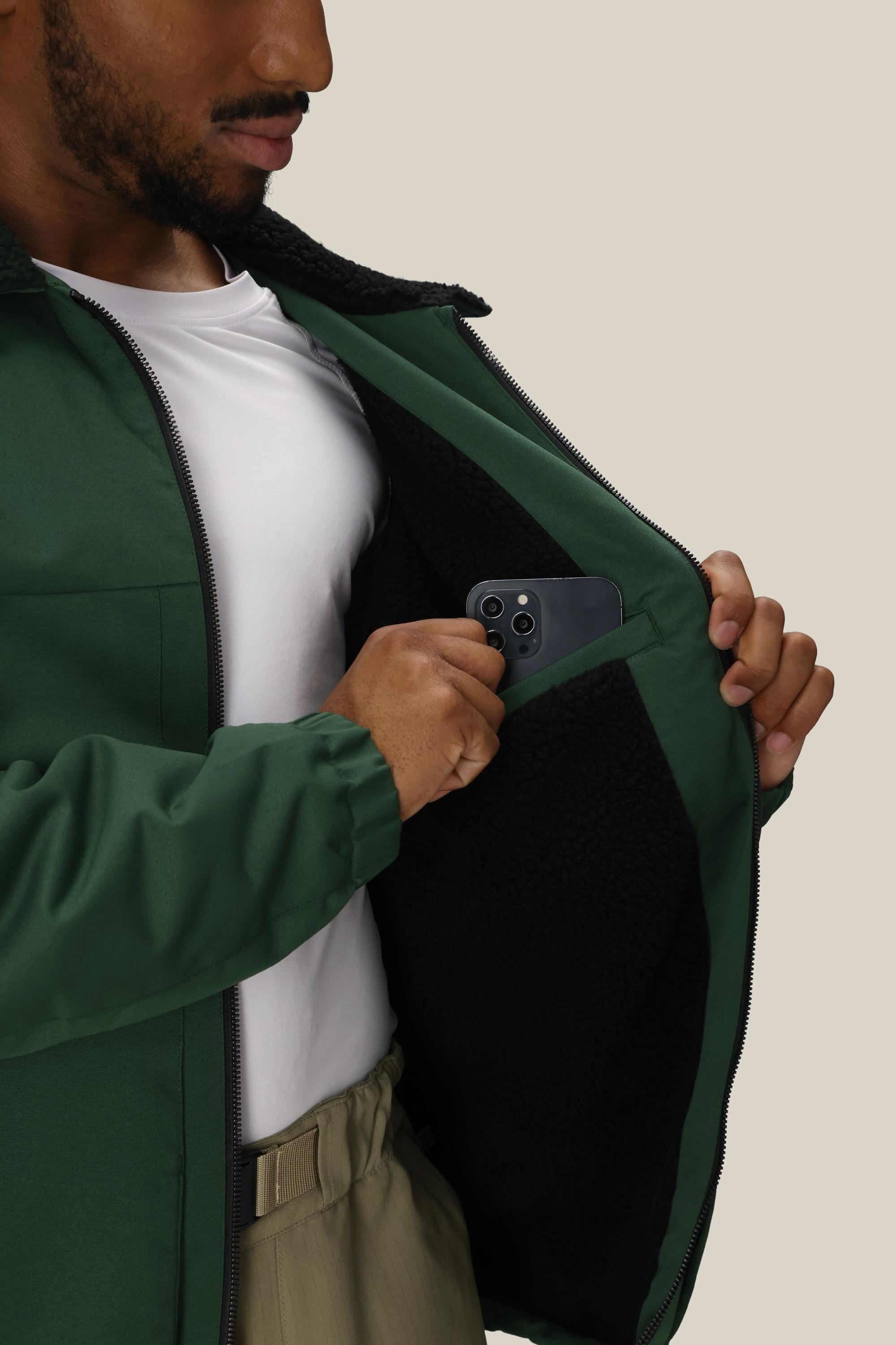 A person wearing the 686 Mens Caim Insulated Jacket partially unzipped, with a white shirt, places a dark smartphone into the inside pocket lined with black fleece. The 686 jacket’s interior pocket is visible.