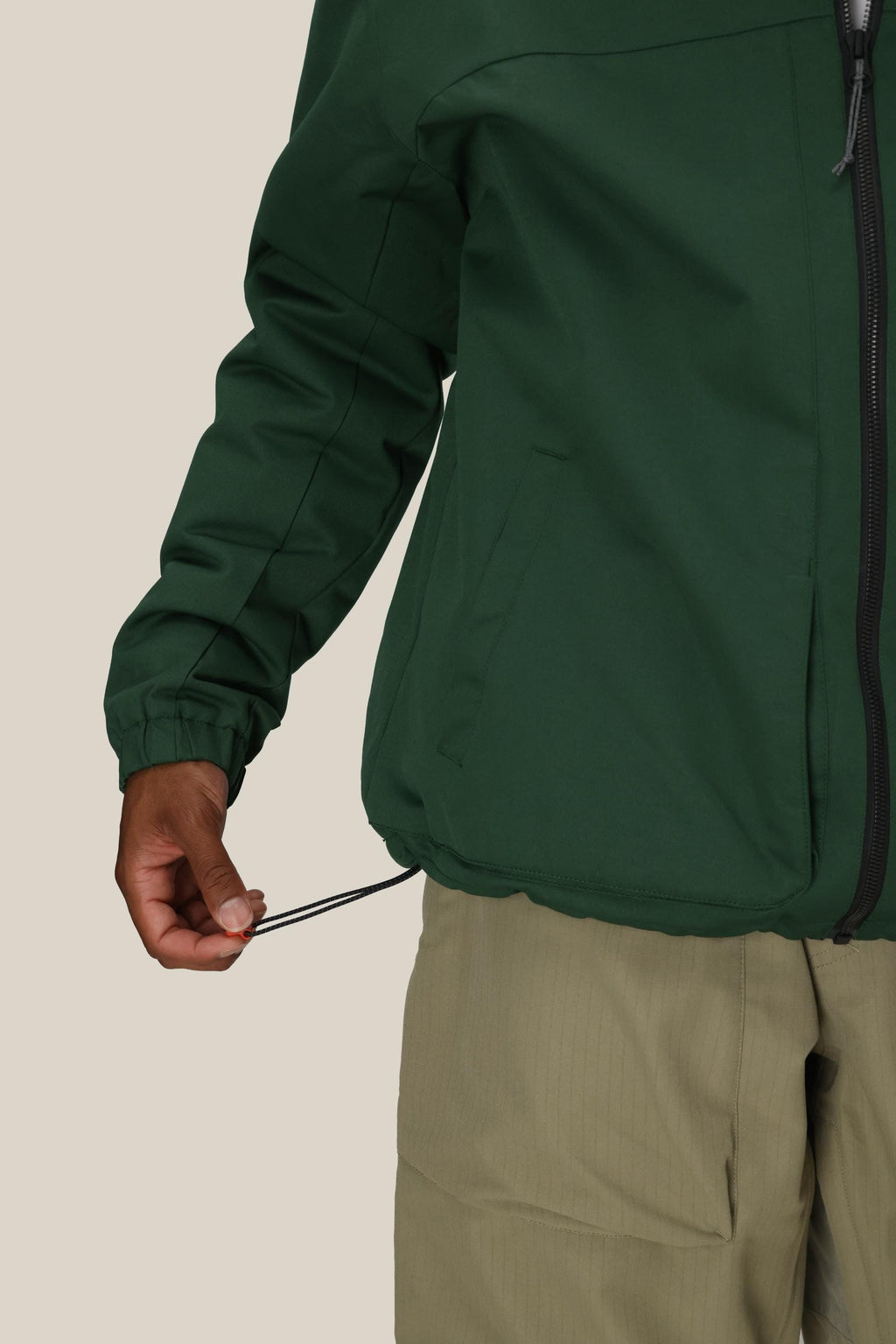 A person wearing the 686 Mens Caim Insulated Jacket in green and khaki pants tightens the jacket’s hem drawstring with one hand against a plain background.