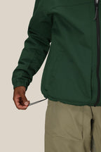 A person wearing the 686 Mens Caim Insulated Jacket in green and khaki pants tightens the jacket’s hem drawstring with one hand against a plain background.