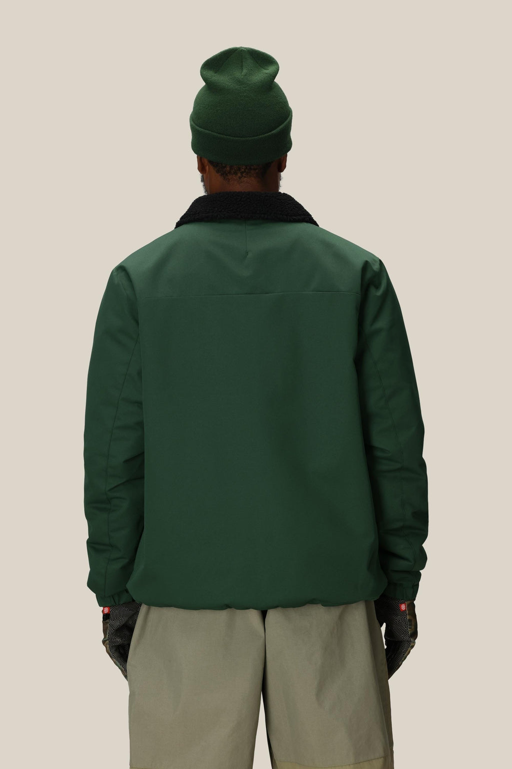 A person faces away against a light beige background, wearing the 686 Mens Caim Insulated Jacket by 686 in green with a black collar, paired with a dark green beanie, gray shorts, and black gloves.