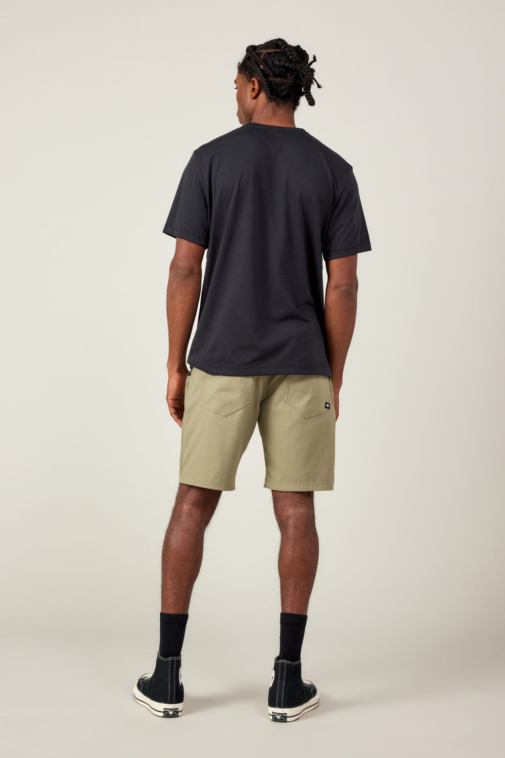 A man stands facing away, wearing a black t-shirt, 686 Men's Everywhere® Ripstop Short in light olive, made with durable stretch fabric, black crew socks, and black sneakers with white soles against a plain light background.
