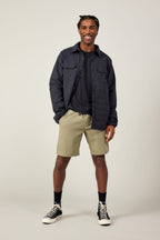 A smiling man stands against a light background wearing a black jacket, dark T-shirt, 686 Men's Everywhere® Ripstop Short by 686, black socks, and black high-top sneakers.
