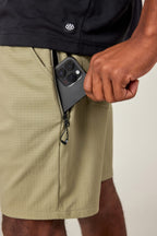 A person places a black smartphone into the front zippered pocket of 686 Men's Everywhere® Ripstop Short in light olive green, paired with a black shirt.