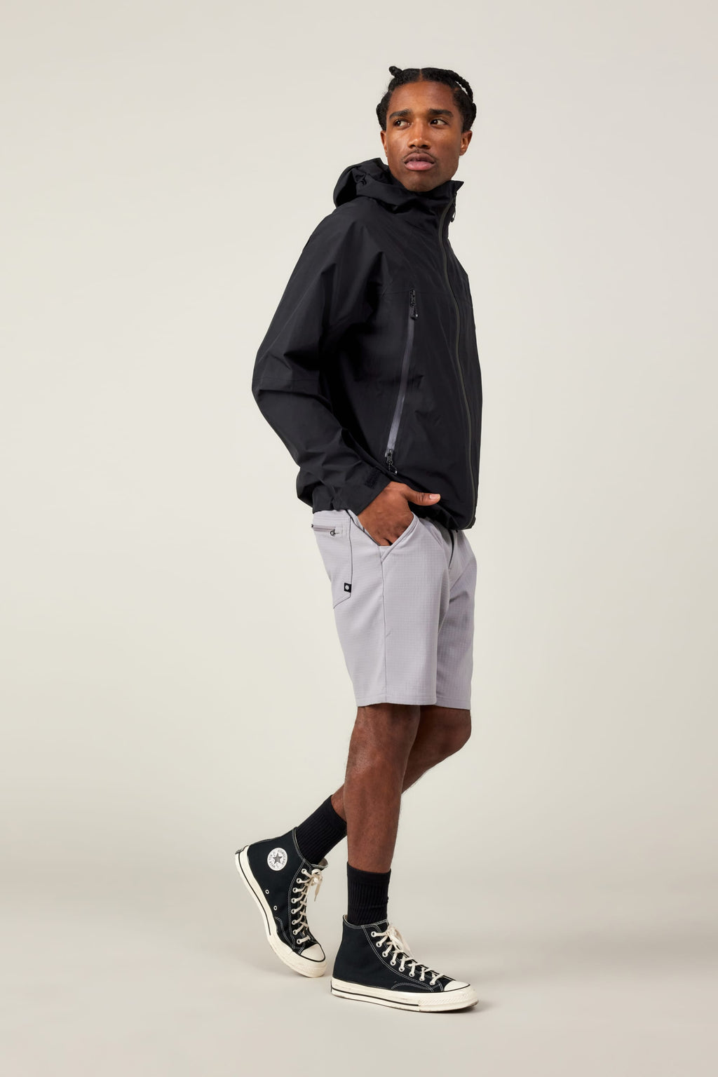 A man stands sideways, looking over his shoulder, hands in his pockets. He wears a black hooded jacket, black socks, black high-top sneakers, and 686 Men's Everywhere® Ripstop Short in light gray against a plain, light background.