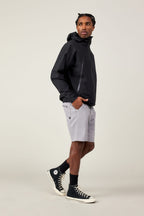 A man stands sideways, looking over his shoulder, hands in his pockets. He wears a black hooded jacket, black socks, black high-top sneakers, and 686 Men's Everywhere® Ripstop Short in light gray against a plain, light background.