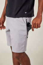 A person wearing 686 Men's Everywhere® Ripstop Short in light gray and a black shirt places a black smartphone into the zippered side pocket of their durable stretch shorts. Only the lower torso and right hand are visible.