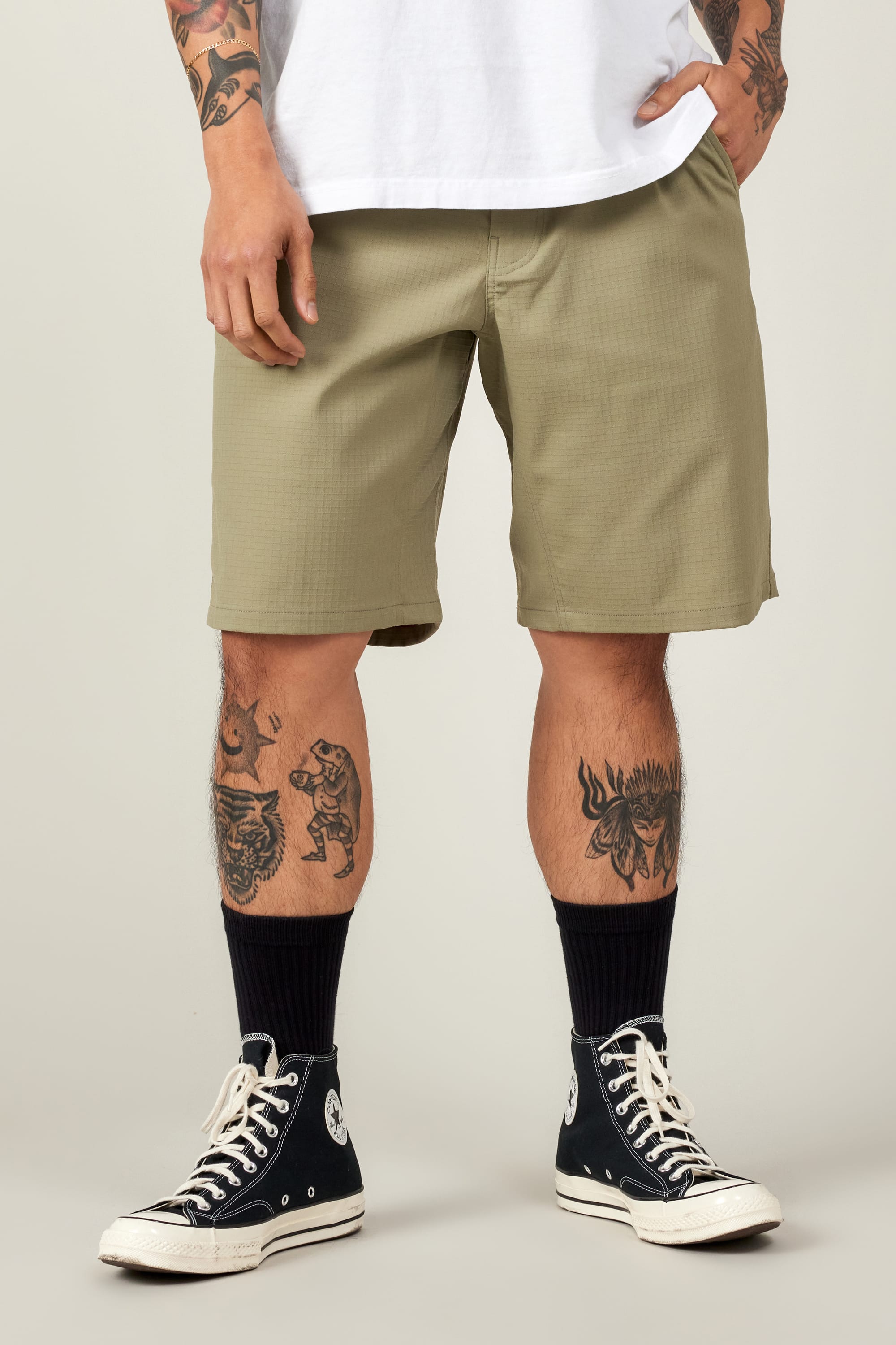 A person wears 686 Men's Everywhere® Ripstop Short - Relaxed Fit in olive green, a white T-shirt, black crew socks, and black high-top Converse. Their tattooed legs and hands rest by their sides against a plain light gray background.