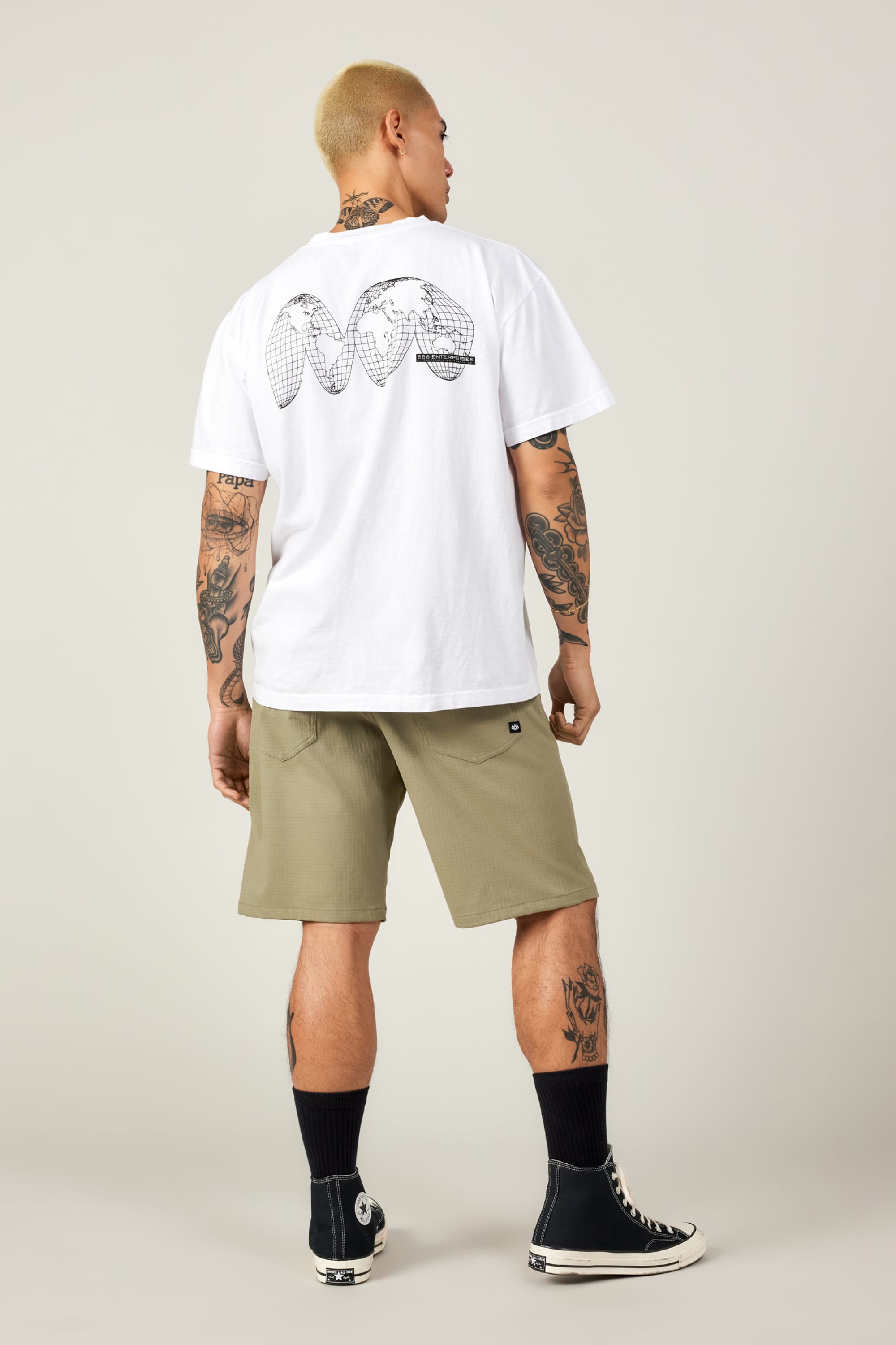 A person with short blond hair and tattoos wears a white graphic tee, black crew socks, black high-top sneakers, and 686 Men’s Everywhere® Ripstop Short - Relaxed Fit, standing with their back to the camera against a plain backdrop.