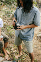 A person with long hair, wearing a light blue shirt and 686 Men's Everywhere® Ripstop Short - Relaxed Fit, stands barefoot outdoors, preparing coffee with a French press. Another barefoot person with a cup sits nearby in a natural, earthy setting.