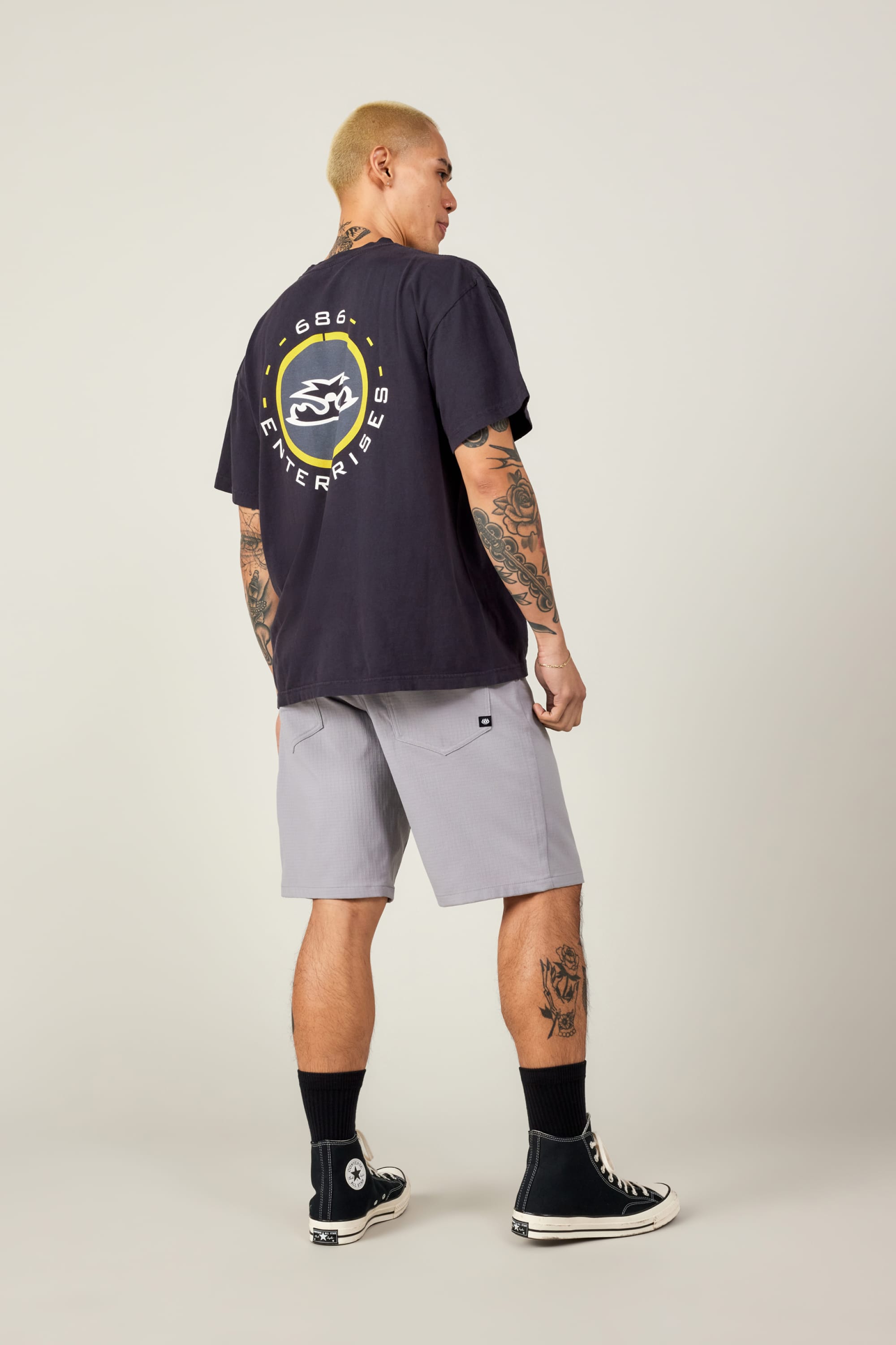 A person with short blond hair and tattoos, in a black graphic tee and 686 Men's Everywhere® Ripstop Short - Relaxed Fit by 686, plus black socks and Converse sneakers, stands facing away against a plain light background.