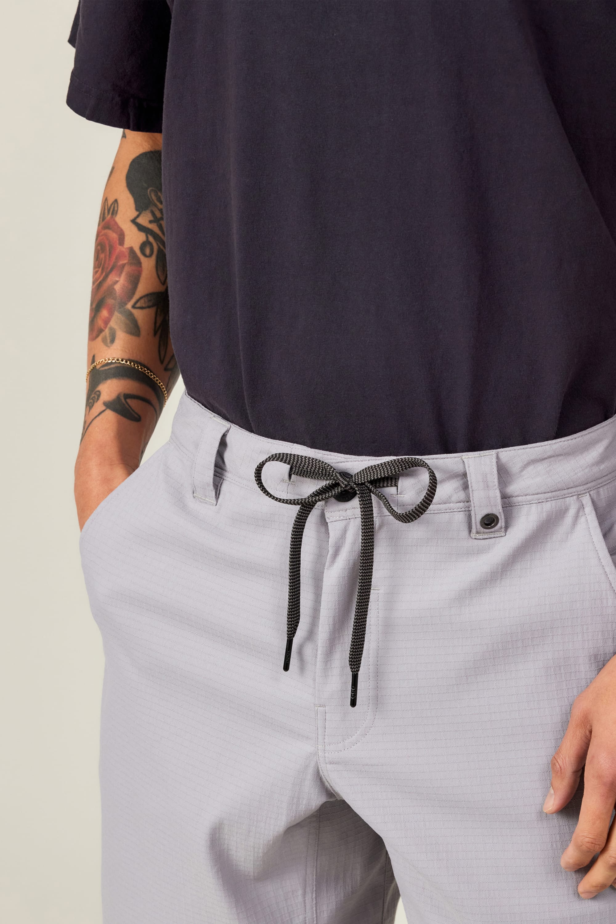 Wearing 686 Men's Everywhere® Ripstop Short - Relaxed Fit with a black drawstring belt and a dark navy shirt, the person stands with hands in pockets, showing a colorful forearm tattoo.