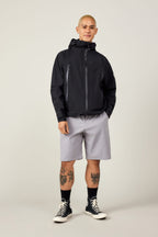 A person with short hair wears a black hooded jacket, 686 Men's Everywhere® Ripstop Short - Relaxed Fit by 686, black high-top sneakers, and shows leg tattoos while standing against a plain light background.