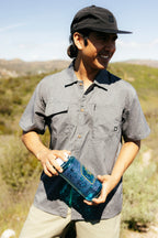 A man wearing the 686 Mens Canopy Perforated Button Up smiles outdoors under a clear sky, holding a blue patterned water bottle with hills and greenery in the background.
