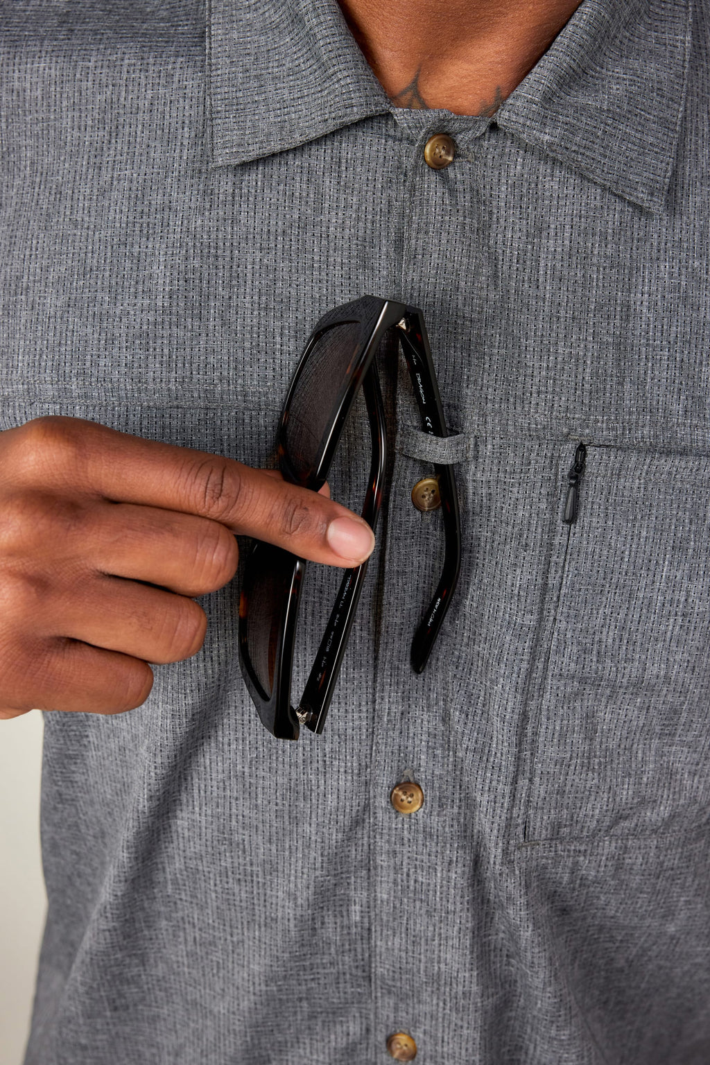 Wearing the 686 Mens Canopy Perforated Button Up in gray, a person slips black sunglasses into a small loop by the chest pocket. Only their torso and hand are shown.