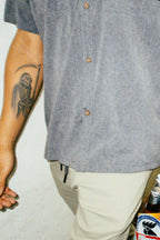 Wearing the 686 Mens Canopy Perforated Button Up in gray and beige pants, a person stands by a wall, showing a Grim Reaper forearm tattoo, with part of a beer bottle visible in the lower right corner.