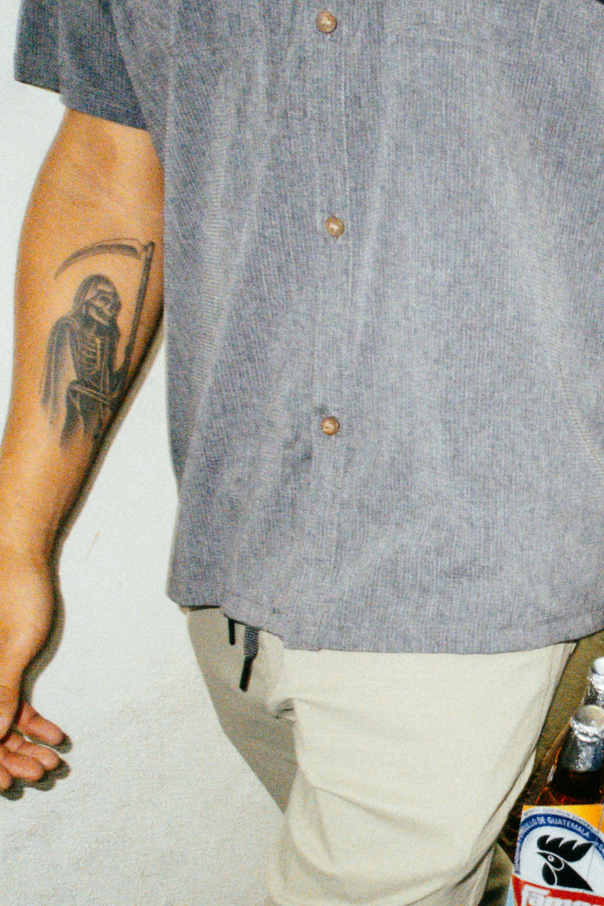 Wearing the 686 Mens Canopy Perforated Button Up in gray and beige pants, a person stands by a wall, showing a Grim Reaper forearm tattoo, with part of a beer bottle visible in the lower right corner.