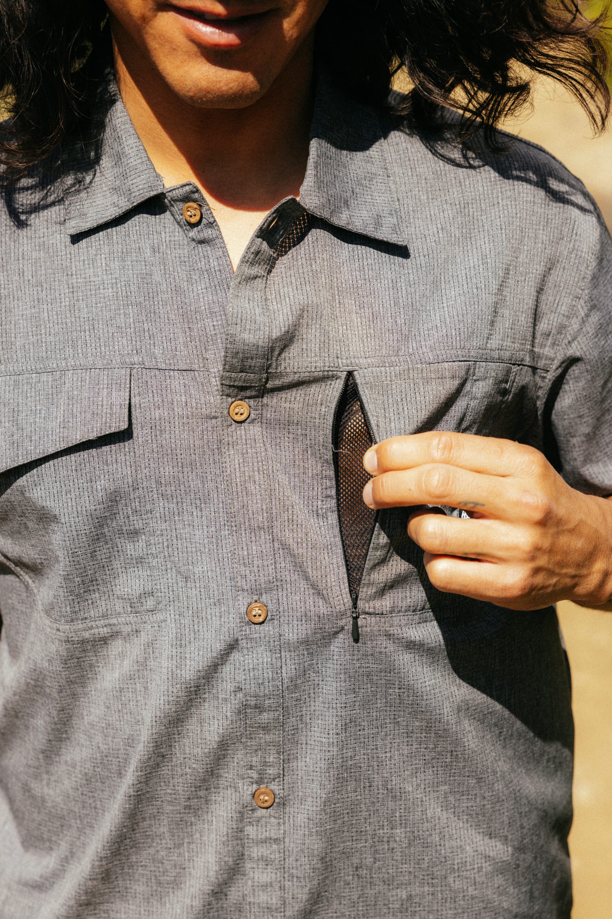 A person with long hair is wearing the 686 Mens Canopy Perforated Button Up and is unzipping a small mesh chest pocket.