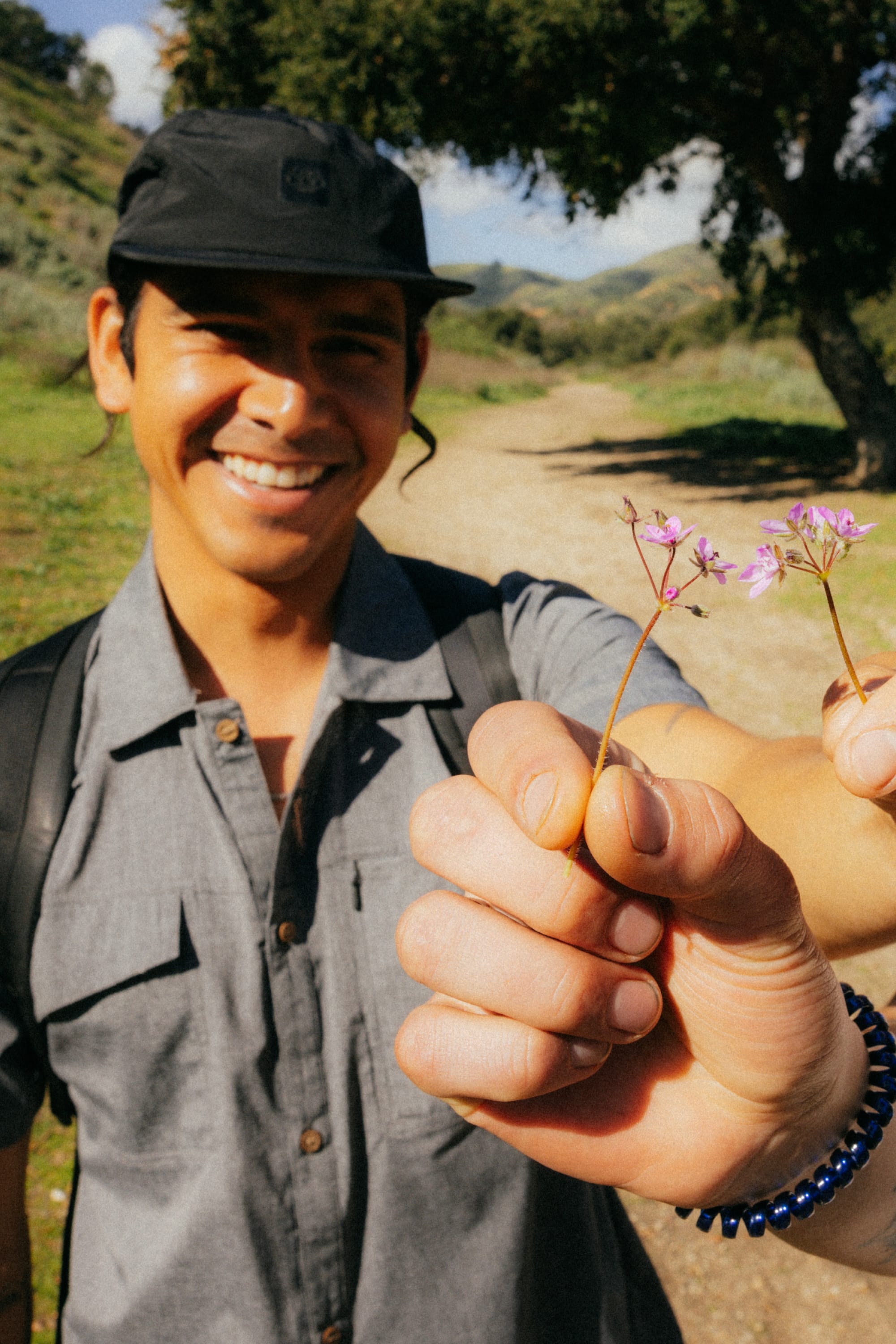 A smiling person outdoors holds two small purple wildflowers toward the camera, wearing the 686 Mens Canopy Perforated Button Up from 686, with a dirt path and green trees in the background.