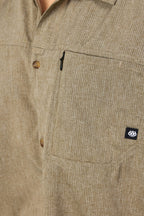 Close-up of the 686 Mens Canopy Perforated Button Up, a beige moisture-wicking shirt with subtle vertical stripes, zippered chest pocket, and a small black 686 logo tag.