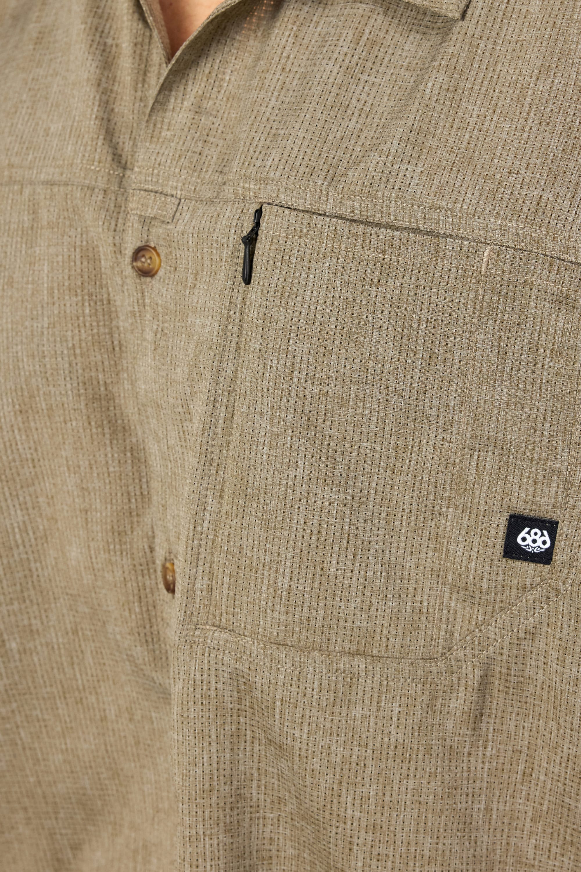 Close-up of the 686 Mens Canopy Perforated Button Up, a beige moisture-wicking shirt with subtle vertical stripes, zippered chest pocket, and a small black 686 logo tag.