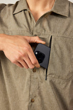 A person wearing the 686 Mens Canopy Perforated Button Up in tan puts a black smartphone with visible cameras into its zippered chest pocket.