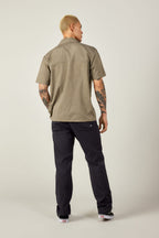 A person with short blond hair and arm tattoos faces away, wearing the 686 Mens Canopy Perforated Button Up in beige, paired with dark pants and sneakers, set against a plain light background.