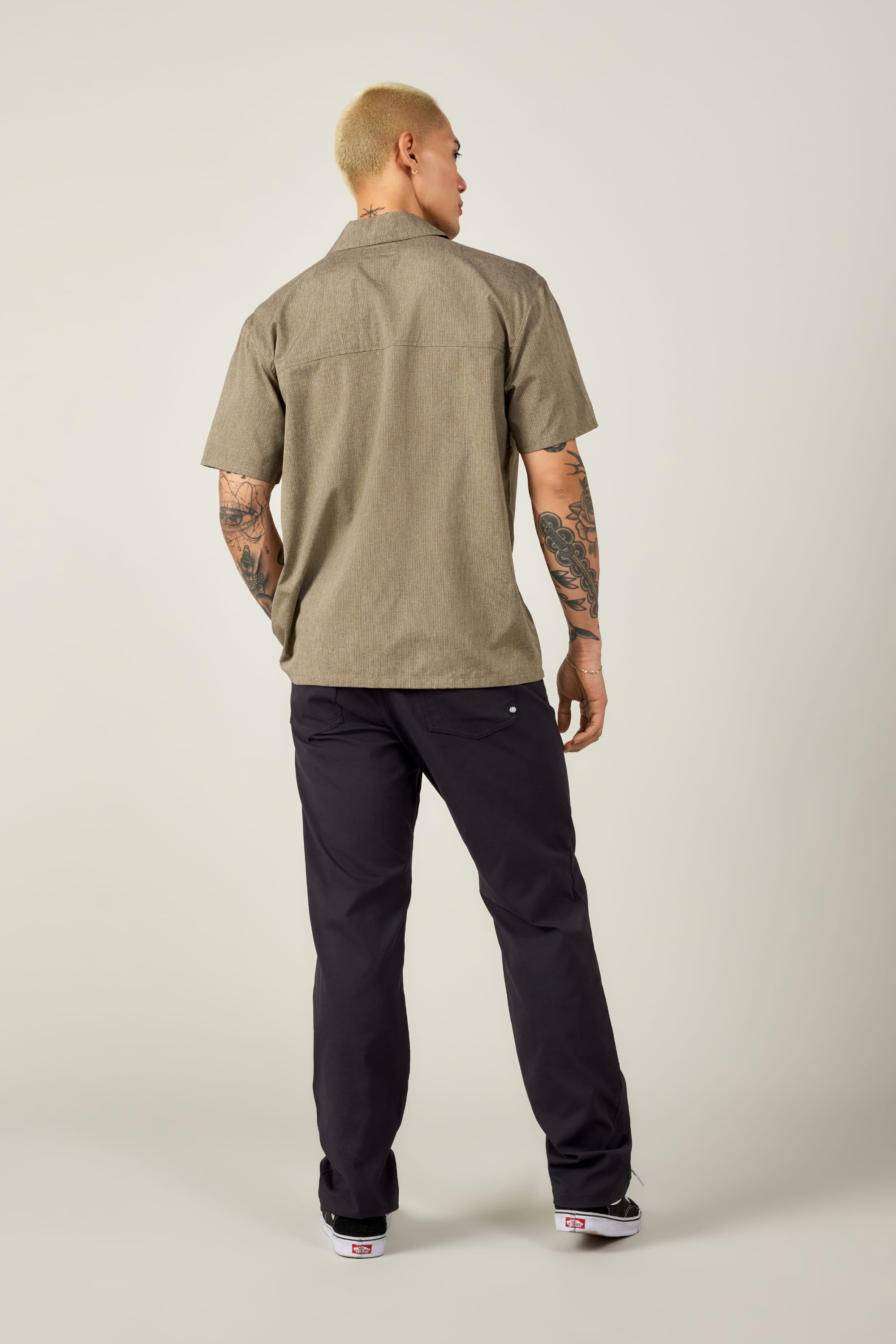 A person with short blond hair and arm tattoos faces away, wearing the 686 Mens Canopy Perforated Button Up in beige, paired with dark pants and sneakers, set against a plain light background.
