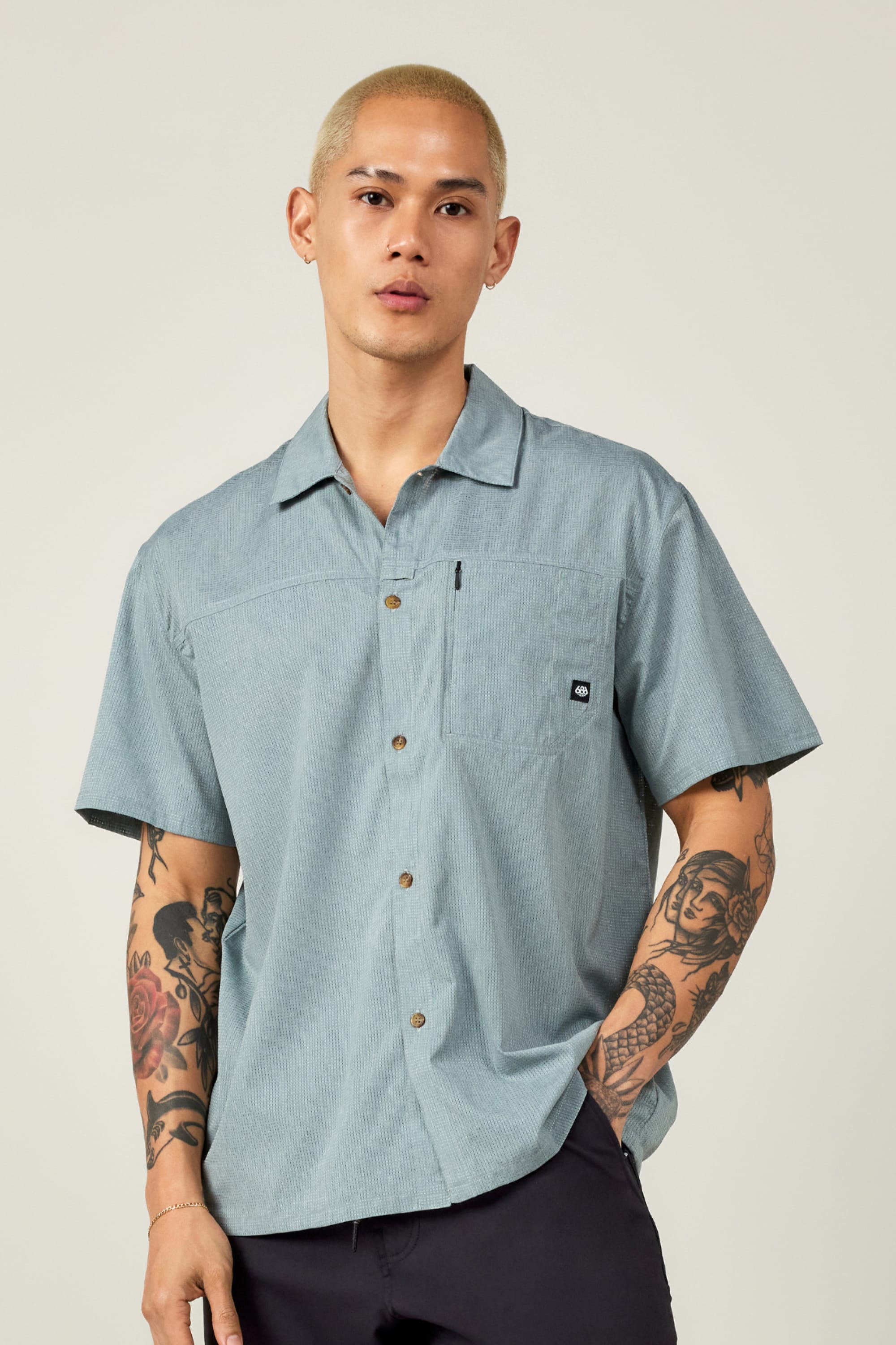 A person with short bleached blond hair and arm tattoos wears the 686 Mens Canopy Perforated Button Up—a light blue, moisture-wicking shirt with a small pocket logo—standing against a plain background.