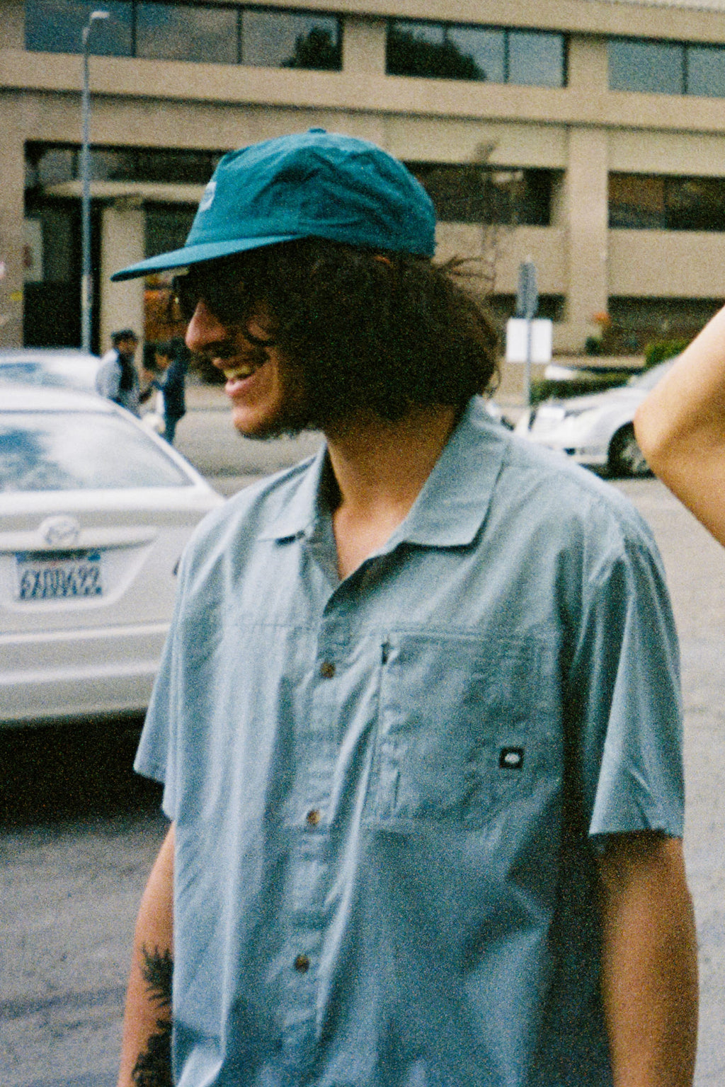A person with long dark hair, wearing a teal cap and a 686 Mens Canopy Perforated Button Up by 686, smiles outdoors near parked cars and a building.