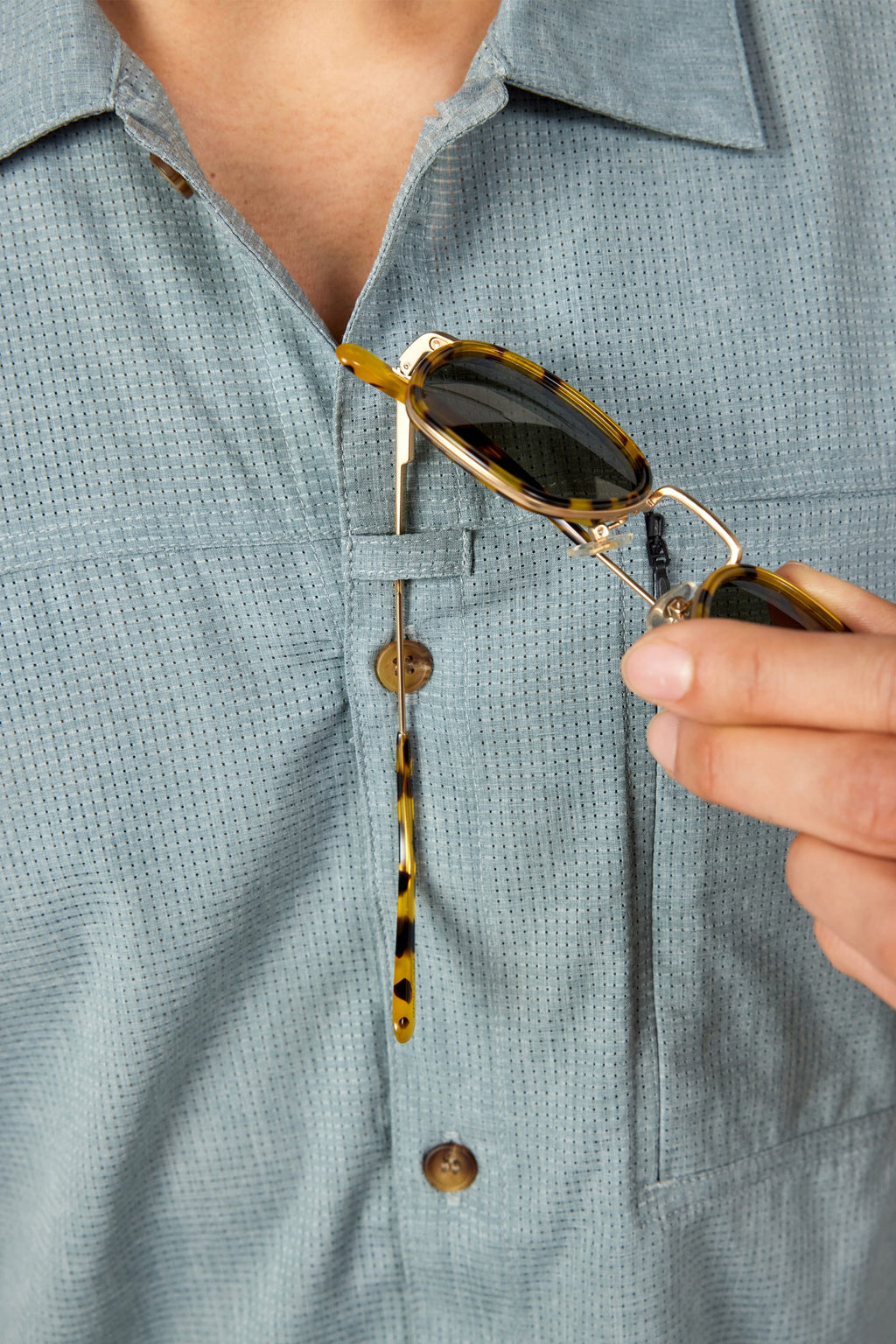 A person wearing the 686 Mens Canopy Perforated Button Up in light blue has tortoiseshell sunglasses hooked onto the shirt’s chest pocket.